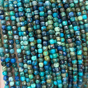 Può includere: Una collana di perline di pietre preziose blu e verdi. Le perline sono sfaccettate e hanno una forma quadrata. Le perline sono infilate su un filo trasparente.