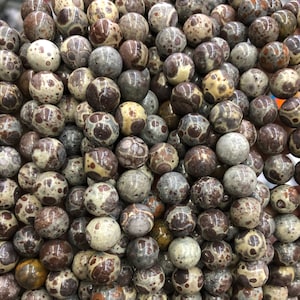 Puede incluir: Primer plano de un montón de cuentas redondas y moteadas. Las cuentas son una mezcla de colores, incluyendo marrón, crema y gris. El patrón moteado les da un aspecto único y natural. Estas cuentas podrían usarse para hacer joyas u otras manualidades.
