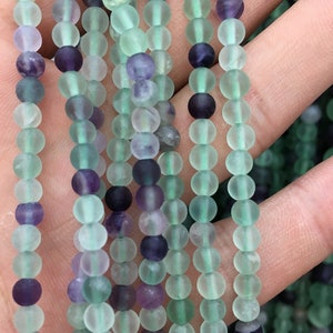 May include: A strand of round, frosted, green and purple fluorite beads. The beads are approximately 6mm in diameter.