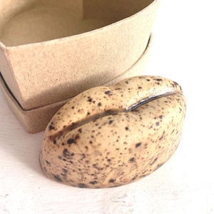 May include: A tan, speckled ceramic coffee bean-shaped object next to a heart-shaped cardboard box. The bean has dark brown spots and a split down the middle. The box is made of light brown cardboard.