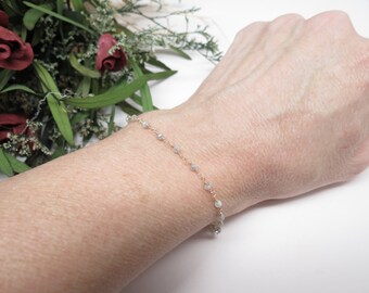 May include: A delicate bracelet with a chain made of small, light gray beads. The bracelet is worn on a person's wrist.