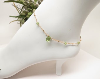 May include: A delicate gold chain anklet featuring small, round, light green beads spaced evenly along the length, with a single, larger, teardrop-shaped light green gemstone charm dangling from the center of the chain.