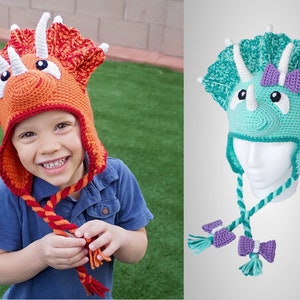 Może przedstawiać: Dwa dzianinowe czapki w kształcie dinozaura, jedna pomarańczowa, a druga turkusowa, z białymi rogami i czarnymi oczami. Pomarańczowa czapka ma białą kokardkę na górze, a turkusowa czapka ma fioletową kokardkę. Obie czapki mają długie sznurki z frędzlami.