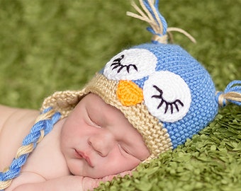May include: A newborn baby wearing a blue and beige crocheted owl hat with white eyes and orange beak. The hat has long beige and blue yarn tails.