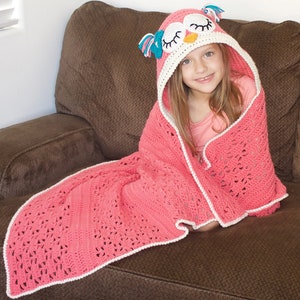 May include: A pink crocheted blanket with a white border and an owl-shaped hood. The hood has a white and orange face with blue eyes and pink accents.
