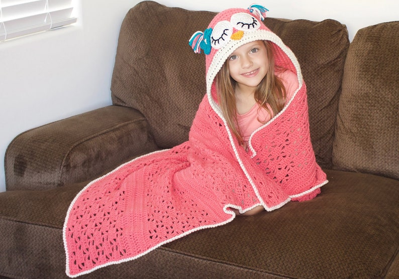May include: A pink crocheted blanket with a white border and an owl-shaped hood. The hood has a white and orange face with blue eyes and pink accents.