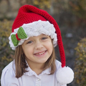 K&ouml;nnte beinhalten: Ein junges M&auml;dchen tr&auml;gt eine rot-wei&szlig;e, geh&auml;kelte Weihnachtsm&uuml;tze mit einer gr&uuml;nen Schleife. Die M&uuml;tze hat einen langen, roten Bommel am Ende.