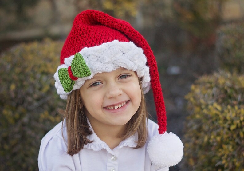 K&ouml;nnte beinhalten: Ein junges M&auml;dchen tr&auml;gt eine rot-wei&szlig;e, geh&auml;kelte Weihnachtsm&uuml;tze mit einer gr&uuml;nen Schleife. Die M&uuml;tze hat einen langen, roten Bommel am Ende.
