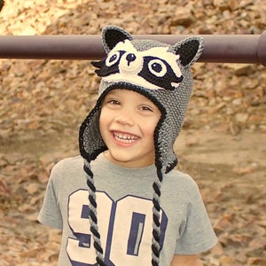 May include: A young person wearing a gray and black raccoon hat with white accents. The hat has black eyes and a black nose. The person is smiling and wearing a gray shirt with a white number 90 on it.