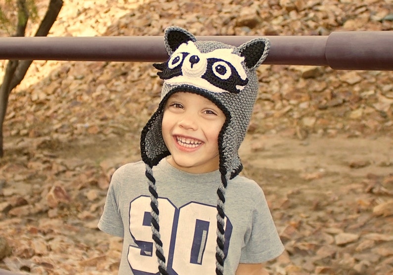May include: A young person wearing a gray and black raccoon hat with white accents. The hat has black eyes and a black nose. The person is smiling and wearing a gray shirt with a white number 90 on it.