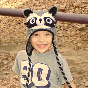 May include: A young child wearing a gray and black raccoon hat with black ear flaps. The hat has white accents and black eyes. The child is wearing a gray shirt with a white number 90 on the front.
