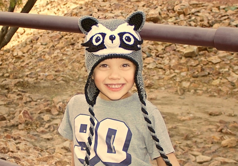 May include: A young child wearing a gray and black raccoon hat with black ear flaps. The hat has white accents and black eyes. The child is wearing a gray shirt with a white number 90 on the front.