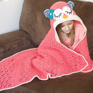 May include: A pink crocheted blanket with a white border and an owl-shaped hood. The hood has a pink, white, and blue owl face with closed eyes.
