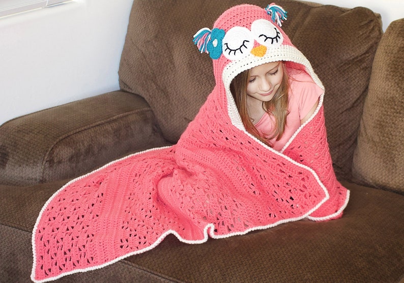 May include: A pink crocheted blanket with a white border and an owl-shaped hood. The hood has a pink, white, and blue owl face with closed eyes.