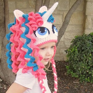 May include: A pink and blue crocheted unicorn hat with a white horn and large blue eyes. The hat has long, curly pink and blue yarn strands that hang down the back.
