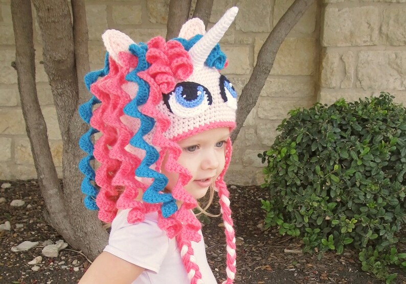 May include: A pink and blue crocheted unicorn hat with a white horn and large blue eyes. The hat has long, curly pink and blue yarn strands that hang down the back.