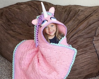 May include: A pink crocheted unicorn blanket with a light blue and purple trim. A young girl is wearing the blanket as a hooded cape.