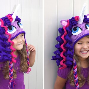 May include: A purple and pink crocheted unicorn hat with a white horn and blue eyes. The hat has long, braided strands of yarn that hang down the back.