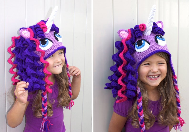 May include: A purple and pink crocheted unicorn hat with a white horn and blue eyes. The hat has long, braided strands of yarn that hang down the back.