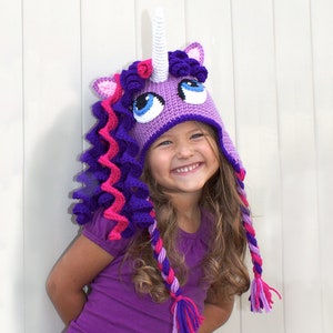 May include: A young girl wearing a purple and pink crocheted unicorn hat with a white horn. The hat has a curly mane and tail.