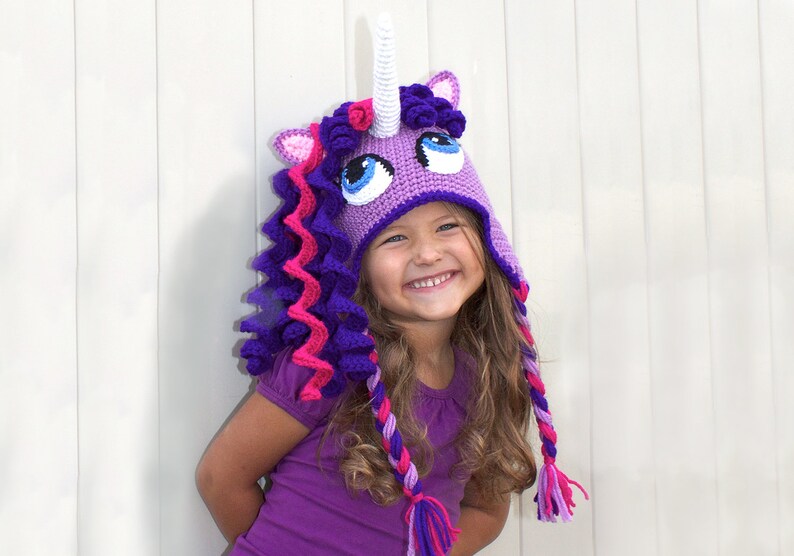 May include: A young girl wearing a purple and pink crocheted unicorn hat with a white horn. The hat has a curly mane and tail.