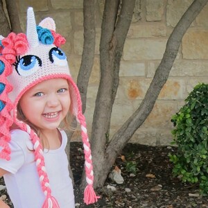May include: A young child wearing a pink and blue crocheted unicorn hat with a white horn and long braids. The hat has large blue eyes and a pink and white braided tail.