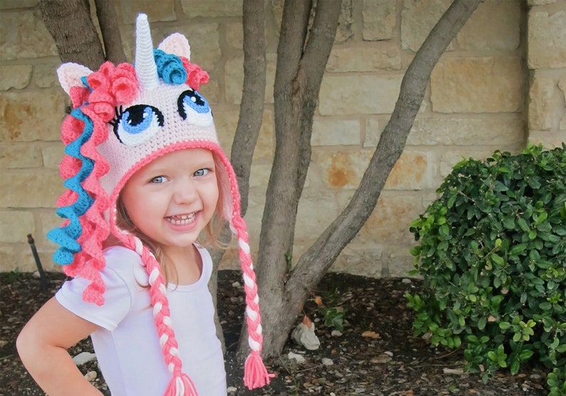 May include: A young child wearing a pink and blue crocheted unicorn hat with a white horn and long braids. The hat has large blue eyes and a pink and white braided tail.
