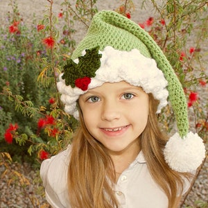 K&ouml;nnte beinhalten: Ein junges M&auml;dchen tr&auml;gt eine gr&uuml;ne und wei&szlig;e H&auml;kelm&uuml;tze mit einem Weihnachtsbaum-Motiv. Die M&uuml;tze hat einen wei&szlig;en Bommel am Ende der spitzen Spitze.