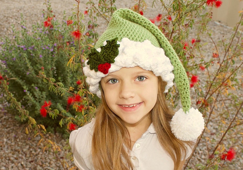 K&ouml;nnte beinhalten: Ein junges M&auml;dchen tr&auml;gt eine gr&uuml;ne und wei&szlig;e H&auml;kelm&uuml;tze mit einem Weihnachtsbaum-Motiv. Die M&uuml;tze hat einen wei&szlig;en Bommel am Ende der spitzen Spitze.