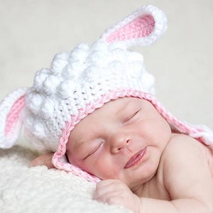 May include: A newborn baby wearing a handmade white and pink crochet lamb hat with long ears. The baby is lying on a soft, white surface, eyes closed, with a slight smile.