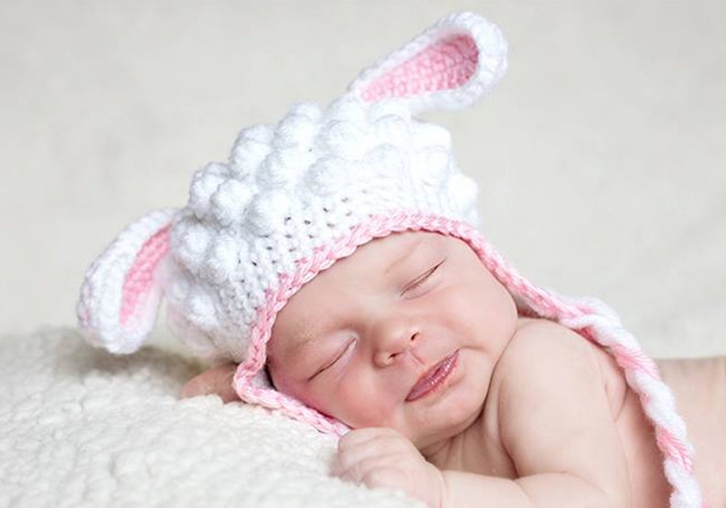 May include: A newborn baby wearing a handmade white and pink crochet lamb hat with long ears. The baby is lying on a soft, white surface, eyes closed, with a slight smile.