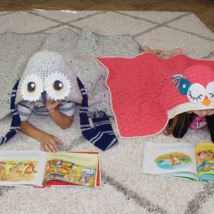 May include: Two children are reading books while wearing crocheted owl hooded blankets. The girl's blanket is pink and the boy's blanket is gray.