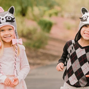 May include: Two children wearing gray raccoon hats with pink and gray braids. The girl's hat has a pink bow. The boy is wearing a black and white diamond patterned sweater.