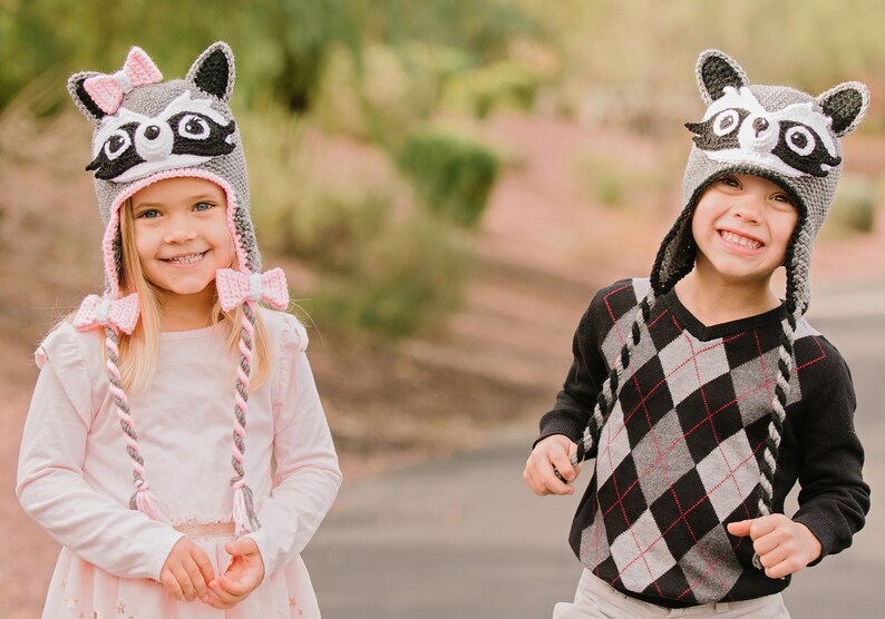 May include: Two children wearing gray raccoon hats with pink and gray braids. The girl's hat has a pink bow. The boy is wearing a black and white diamond patterned sweater.