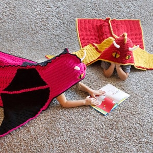 May include: Two children wearing crocheted dragon costumes are lying on a carpet. One child is reading a book titled "Love You"