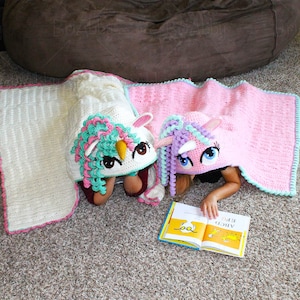 May include: Two children are reading a book while wearing crocheted unicorn hooded blankets. The blankets are pink and white with a unicorn face on the hood. The book is yellow with the words "Are You My Mother?" on the cover.
