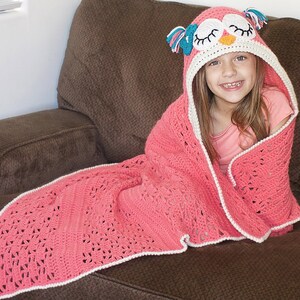 May include: A pink crocheted blanket with a white border and an owl-shaped hood. The hood has a white face with black eyes and a yellow beak. The blanket is draped over a brown couch.