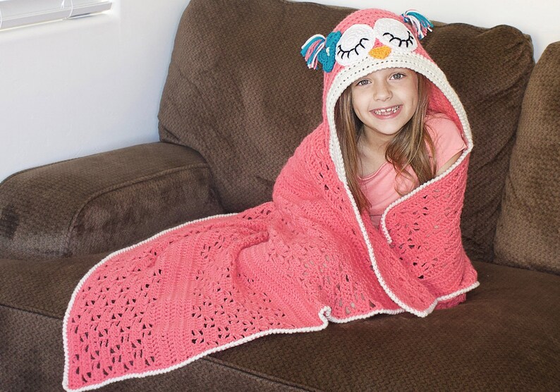 May include: A pink crocheted blanket with a white border and an owl-shaped hood. The hood has a white face with black eyes and a yellow beak. The blanket is draped over a brown couch.