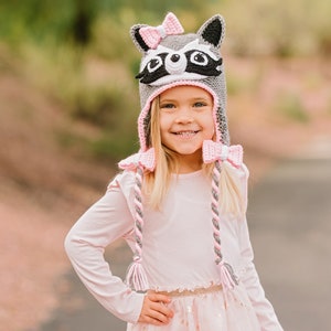 May include: A young girl wearing a gray and pink raccoon hat with a pink bow. The hat has long, braided ties with pink and gray yarn.