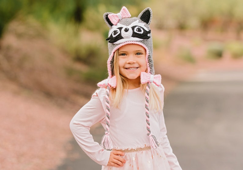 May include: A young girl wearing a gray and pink raccoon hat with a pink bow. The hat has long, braided ties with pink and gray yarn.