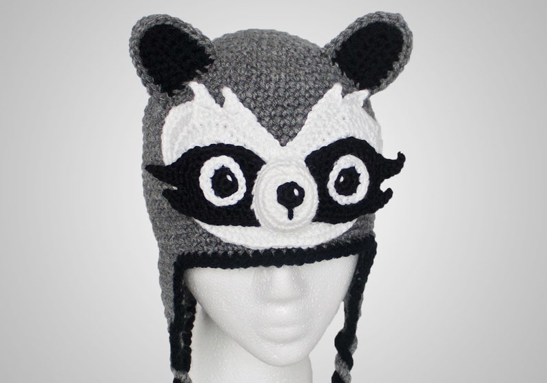 May include: Gray crocheted hat with black and white raccoon face details. The hat has long ear flaps and a black crocheted brim.