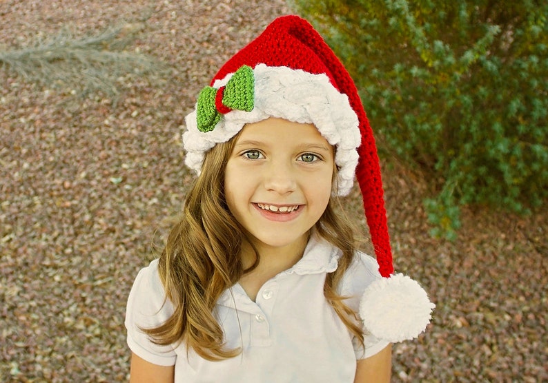 K&ouml;nnte beinhalten: Ein junges M&auml;dchen tr&auml;gt eine rot-wei&szlig;e geh&auml;kelte Weihnachtsm&uuml;tze mit einer gr&uuml;nen Schleife. Die M&uuml;tze hat einen langen, roten Bommel am Ende.