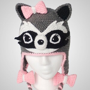 May include: Gray and black crocheted raccoon hat with pink bows on the top and the ties. The hat has a white face with black eyes and a black nose.