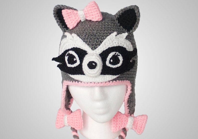 May include: Gray and black crocheted raccoon hat with pink bows on the top and the ties. The hat has a white face with black eyes and a black nose.