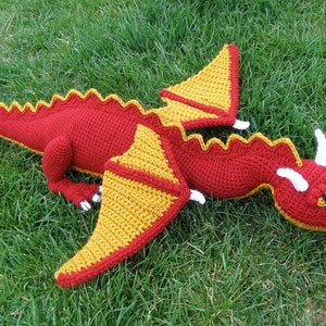 May include: A red and yellow crocheted dragon lying on green grass. The dragon has white horns and a yellow eye.
