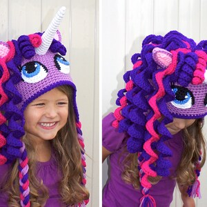 May include: A purple and pink crocheted unicorn hat with a white horn. The hat has a face with blue eyes and a pink nose. The hat is being worn by a child.