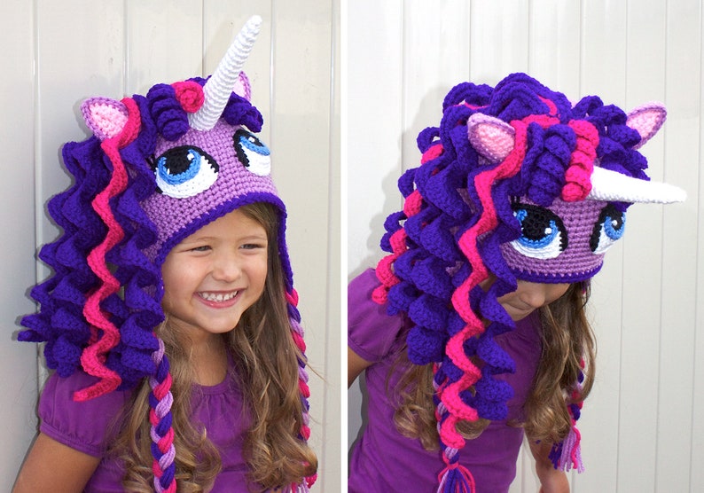 May include: A purple and pink crocheted unicorn hat with a white horn. The hat has a face with blue eyes and a pink nose. The hat is being worn by a child.