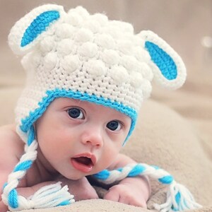 May include: A white and blue crochet baby hat shaped like a sheep, with blue accents on the ears and trim. The hat has a textured, bobble-like design. The hat is worn by a baby.