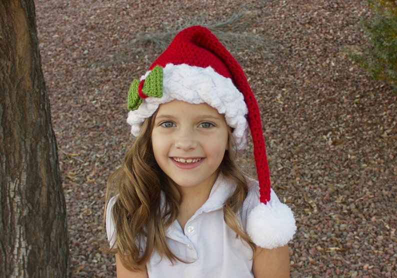 K&ouml;nnte beinhalten: Ein junges M&auml;dchen tr&auml;gt eine rot-wei&szlig;e geh&auml;kelte Weihnachtsm&uuml;tze mit einer gr&uuml;nen Schleife. Die M&uuml;tze hat einen langen, roten Bommel am Ende.