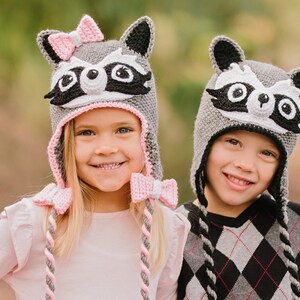 May include: Two children wearing gray and pink raccoon hats with black eyes and noses. The girl's hat has a pink bow on top. The boy's hat has a gray and black braided cord.